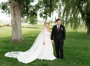Bride and groom stand together on lush green grass beneath a large tree on their wedding day.