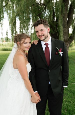 A bride and groom pose together outdoors on their wedding day beneath a willow tree on a sunny day.