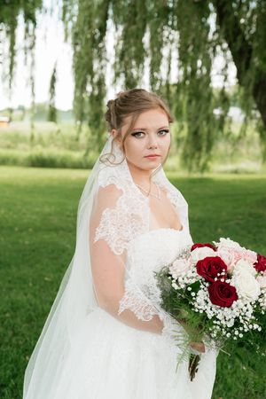 Bride in white lace wedding dress and veil holds red and pink bouquet outdoors near weeping willow tree.