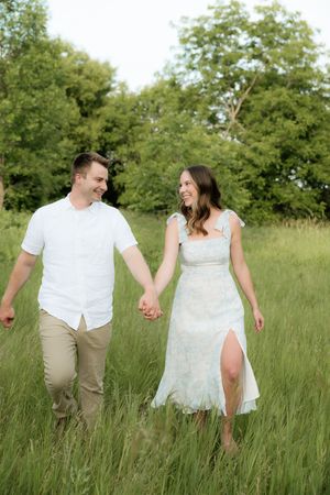 Happy couple holding hands walking through a lush green meadow, man in white shirt and woman in lace dress.