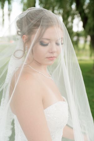 Elegant bride in white strapless gown and pearl necklace, draped in sheer veil, looking down in lush green park.