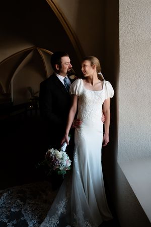 Bride in white lace gown holds bouquet beside groom in black suit, posing in elegant arched interior.