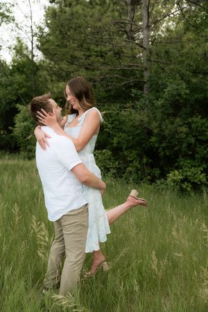 A couple embraces in a lush green meadow, the man lifting the woman in a light floral dress outdoors.