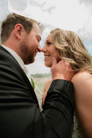 A bride and groom share an intimate moment under a veil, with the groom tenderly holding her face as she smiles.