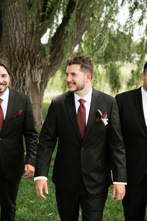 Groomsmen in black suits with burgundy ties and boutonnieres walking outdoors near green trees at a wedding.