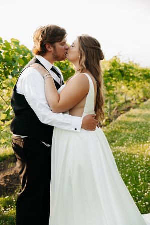 A bride and groom share a romantic kiss in a lush vineyard, she in a white gown, he in a black vest.
