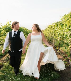 Newlywed couple walking hand in hand through a lush vineyard, bride in white gown and groom in black vest.