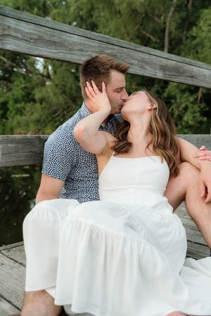 Couple kissing on wooden dock, woman in white dress seated on man in blue patterned shirt outdoors.