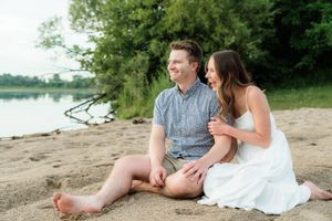 Couple sitting on sandy beach by river, woman in white dress leaning on smiling man in blue shirt.