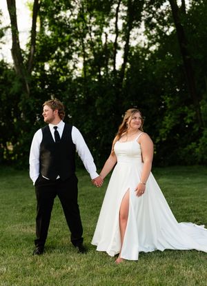 A bride in a white gown and groom in black vest hold hands on green lawn with trees behind them.
