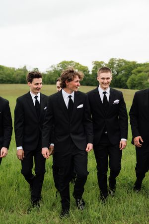 Groomsmen in black suits with white pocket squares walk across a green field on a wedding day.