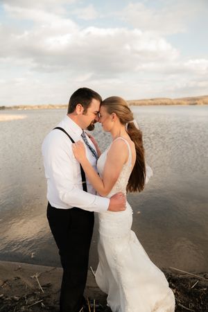 A bride and groom share a romantic forehead touch by a serene lake during golden hour in their wedding attire.
