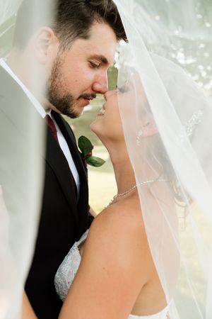 Bride and groom share an intimate moment under a flowing white veil, nearly kissing in a romantic outdoor wedding portrait.
