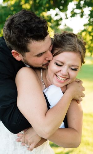 Groom hugs and kisses smiling bride outdoors in green garden setting.