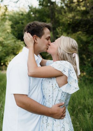 A couple shares a romantic kiss outdoors surrounded by lush green trees during their engagement photo session.