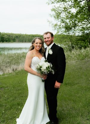 Smiling bride and groom pose outdoors near a lake, bride holding white bouquet in elegant strapless wedding gown.