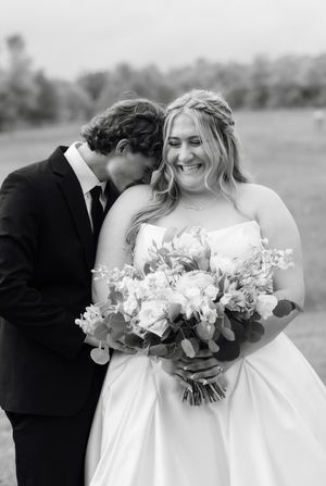 A smiling bride holds a floral bouquet as her groom nuzzles her cheek in a black and white wedding photo.