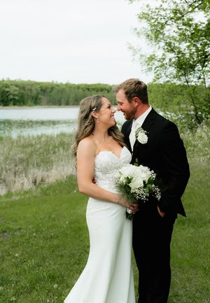 A bride and groom share a romantic moment outdoors near a lake, surrounded by lush green trees and grass.