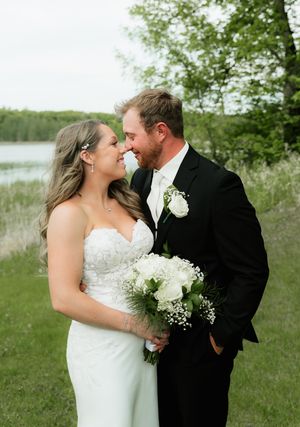 Smiling bride and groom stand nose-to-nose outdoors by a lake, bride holding a white bouquet of roses and baby's breath.