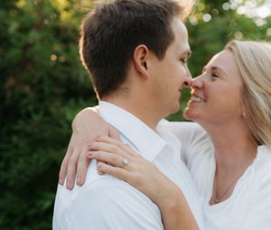 A couple in white embraces and smiles at each other outdoors, with the woman wearing an engagement ring.