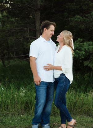 Happy couple in casual white tops and jeans laugh together during an outdoor engagement photo session near pine trees.