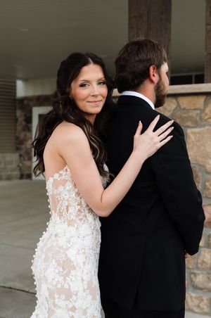 Bride in lace gown embraces groom in black tuxedo from behind, smiling softly at camera during wedding portrait.