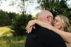 A couple shares a romantic kiss outdoors, woman with blonde hair and visible tattoo embracing a bald man in black shirt.