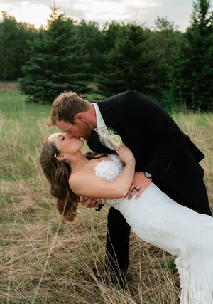 A groom dips his bride for a romantic kiss in a golden field with evergreen trees in the background.