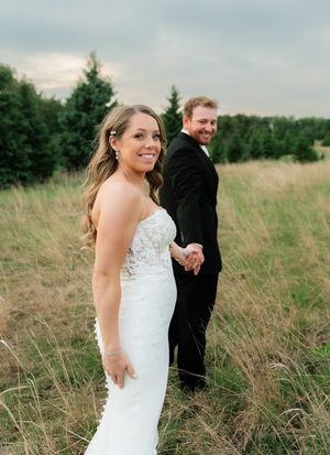 Bride in white lace gown holds grooms hand in golden field with evergreen trees backdrop.