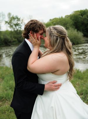 A bride and groom share a romantic moment near a serene pond, dressed in wedding attire outdoors.