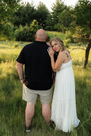 Couple posing outdoors in green field, woman in white dress leaning on man in black shirt, golden hour lighting.