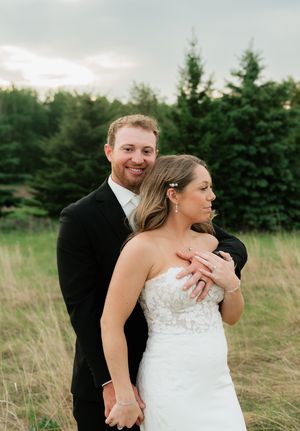A smiling groom in a black tuxedo embraces his bride in a strapless lace wedding gown in a scenic outdoor field.