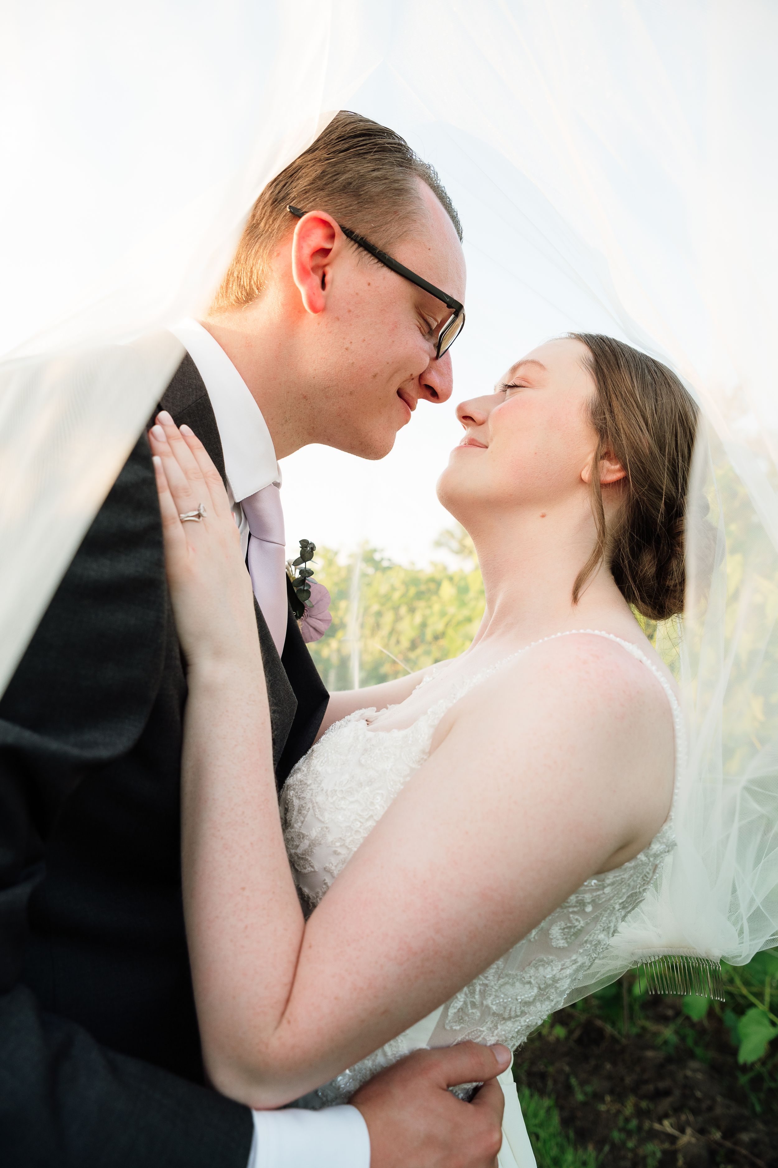 Bride and groom share a romantic moment under a flowing veil, smiling at each other in wedding attire outdoors.