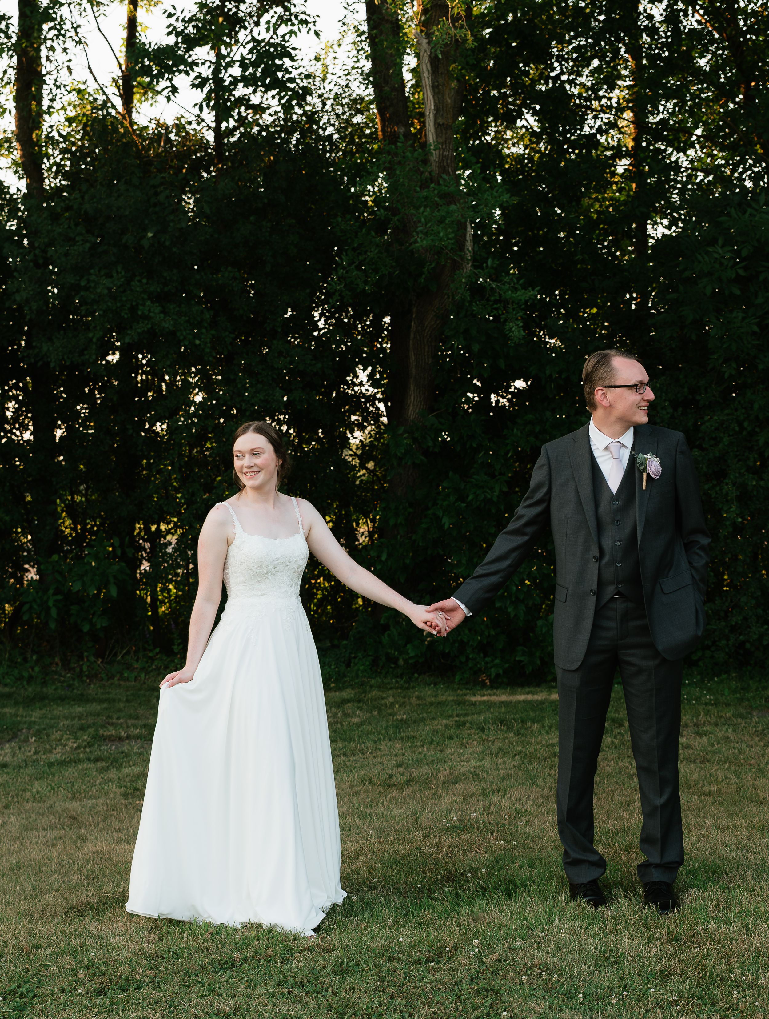 A bride and groom hold hands outdoors on a grassy lawn with lush green trees in the background at golden hour.