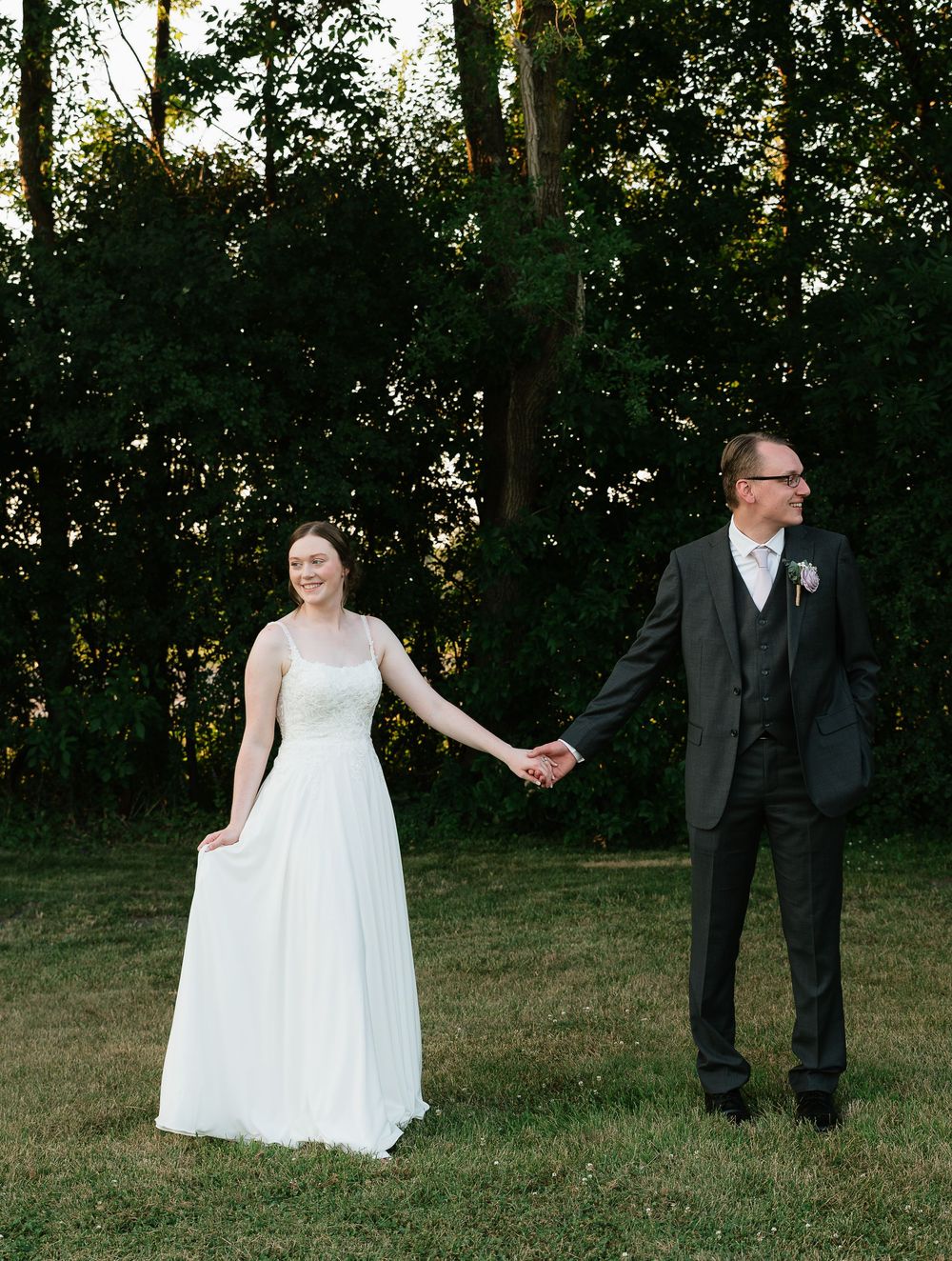 A bride and groom hold hands outdoors on a grassy lawn with lush green trees in the background at golden hour.