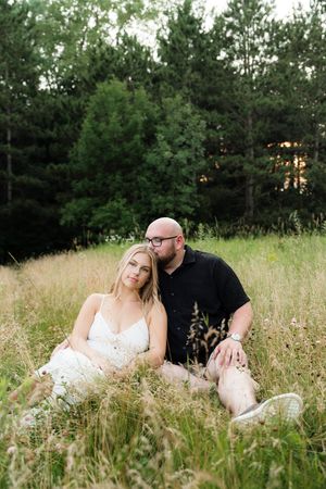 Couple sitting together in tall grass field at sunset with pine trees, woman leaning on mans shoulder.