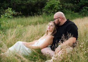 Couple sitting in tall grass field, woman in white dress leaning against man in black shirt, surrounded by lush greenery.