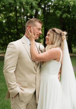 A bride in a white gown and veil tenderly cups a groom's face in a romantic outdoor wedding portrait.