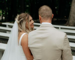 Bride in white gown and veil smiles at groom in beige suit during outdoor wedding ceremony near white benches.