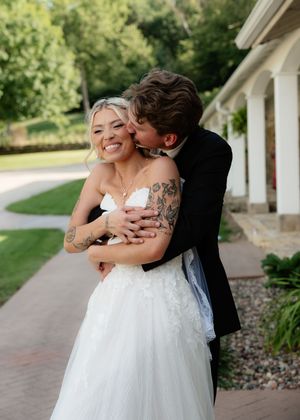 A smiling bride in a white tulle gown with tattoos is embraced and kissed by a groom in a black suit outdoors.