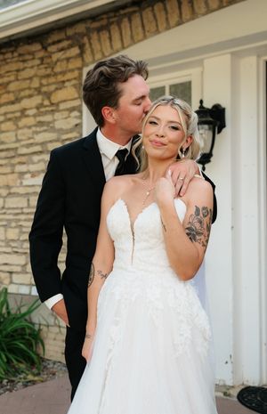 Bride in white lace gown and groom in black suit share a tender moment outside a stone building.