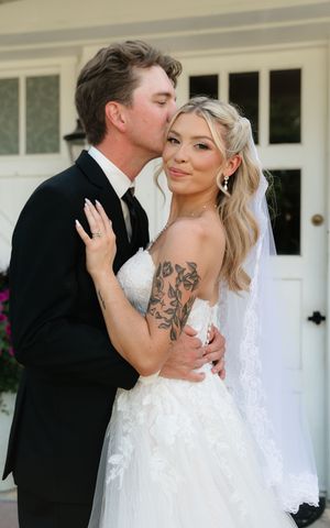 Bride in white strapless gown with veil and floral tattoo poses with groom in black tuxedo kissing her cheek outdoors.
