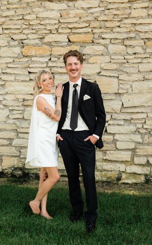 Couple posing by stone wall, woman in white mini dress with tattoos, man in black suit with tie, standing on green grass.