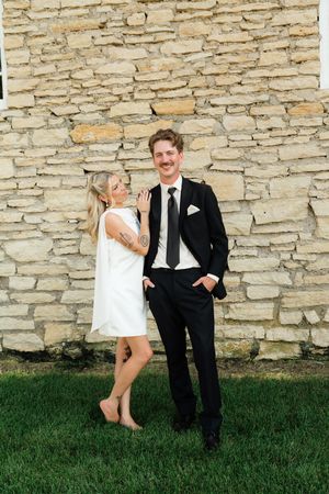 A smiling couple poses against a stone wall, woman in white dress and man in black suit with tie on green grass.