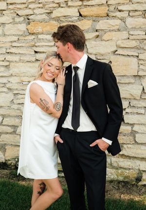 Couple posing by stone wall, woman in white mini dress with tattoos, man in black suit with tie kissing her temple.