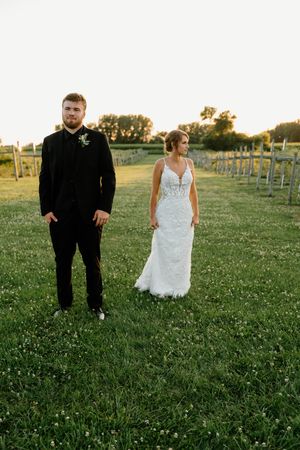 A wedding couple stands in an open field at sunset with vineyard rows in the background.