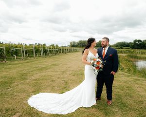 Newlyweds pose together on a path alongside vineyard rows under cloudy skies.