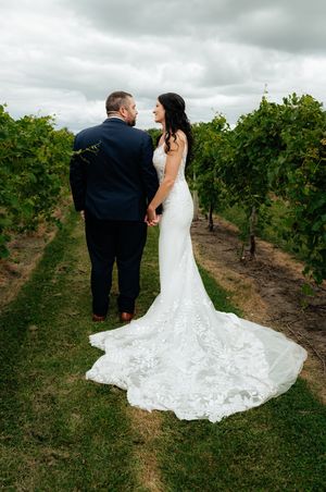 A romantic wedding couple stands between rows of grapevines in a vineyard on an overcast day.
