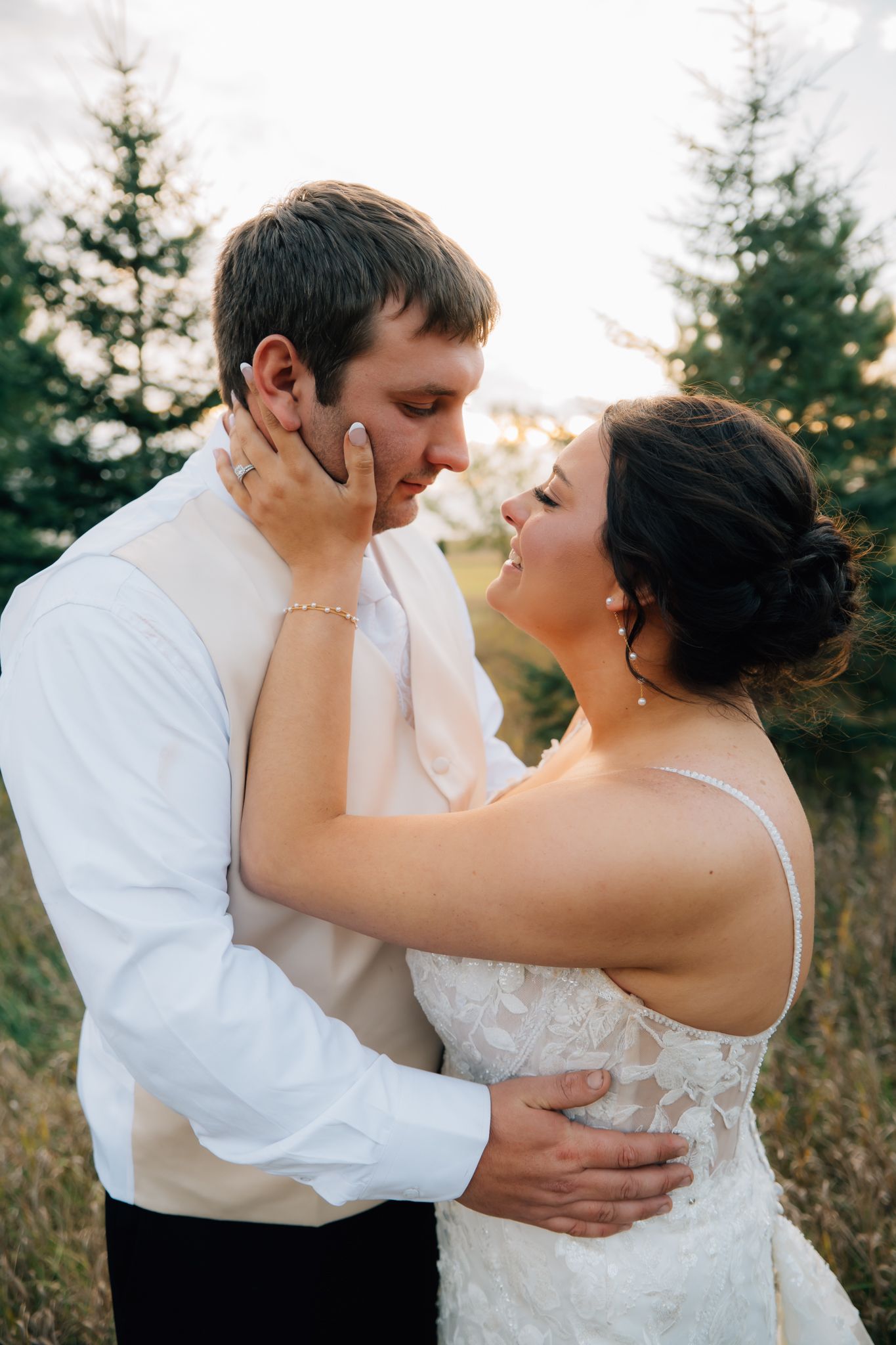 A romantic outdoor wedding portrait of a couple embracing in front of evergreen trees at sunset.