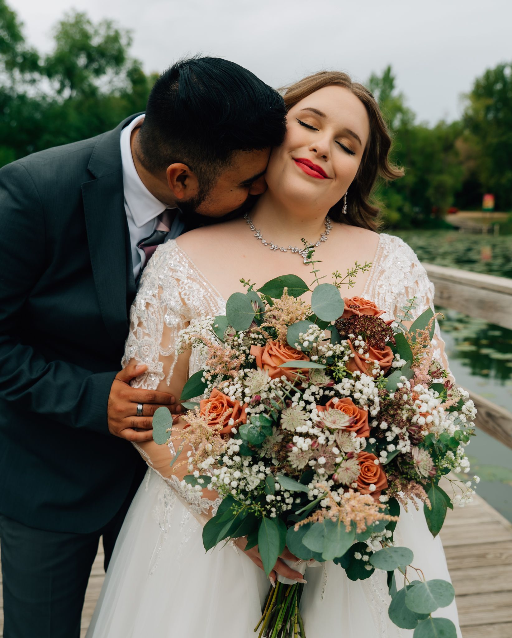 A romantic wedding couple shares tender moments with peach and eucalyptus bouquet on a wooden dock by the water.